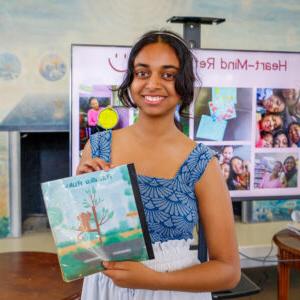 Pratya Poosala holds up her children’s book titled Rajulko Rukh. The book cover is a digital illustration of a brown monkey sitting on a tree by a street. Poosala has dark wavy hair pulled back and wears a blue top and white pants.